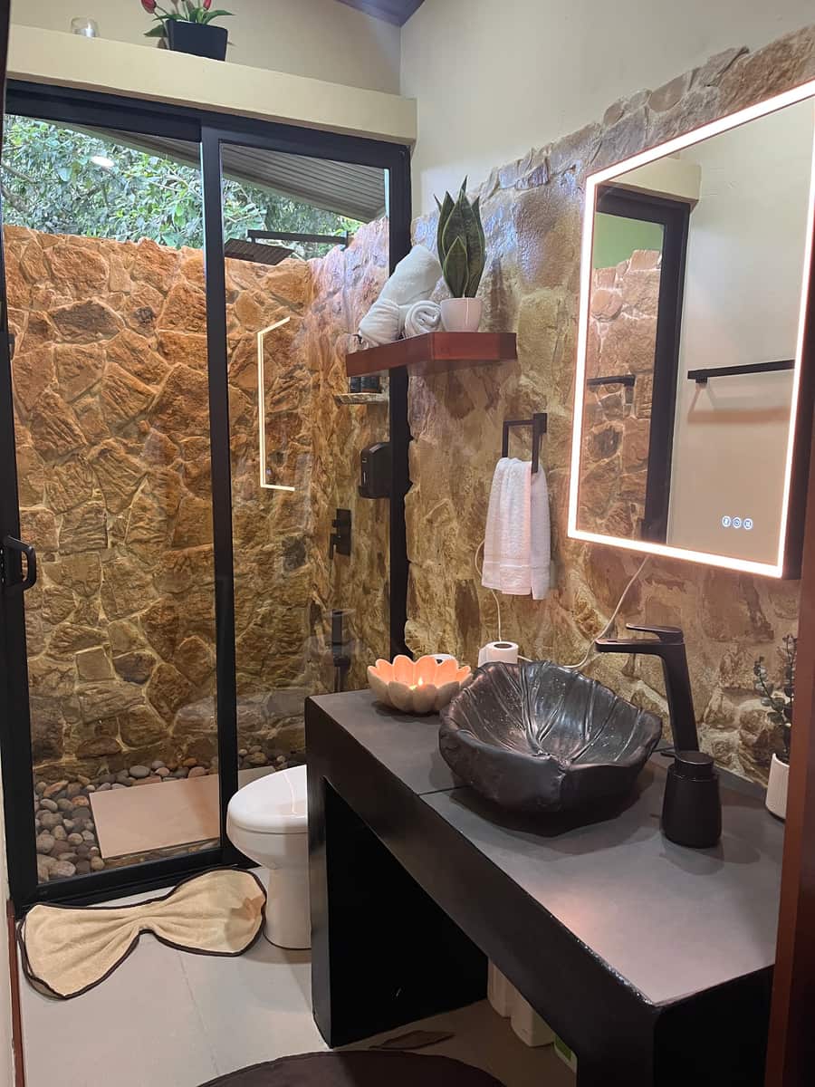 Baño de Reborn Spa con muro de piedra, mampara de vidrio, espejo retroiluminado y lavabo de piedra tallada